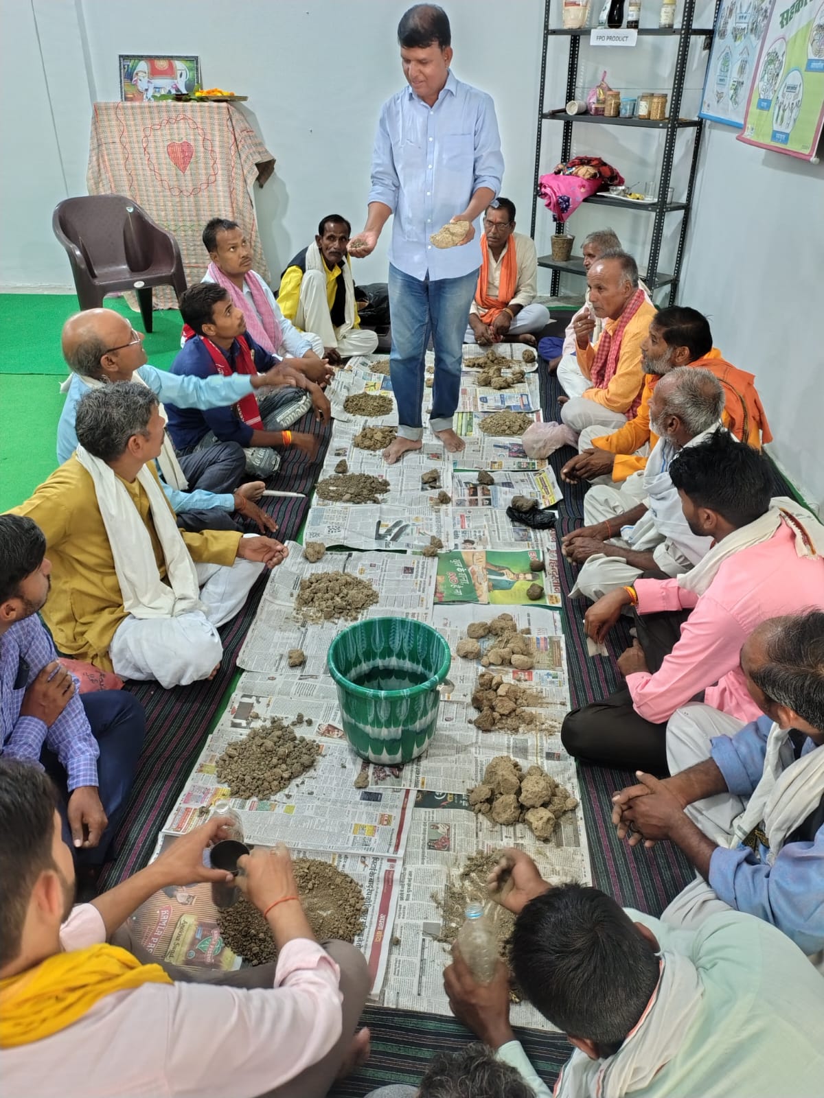 An instructor demonstrating soil testing to farmers seated on the floor around soil samples at the FPO office in Atarra