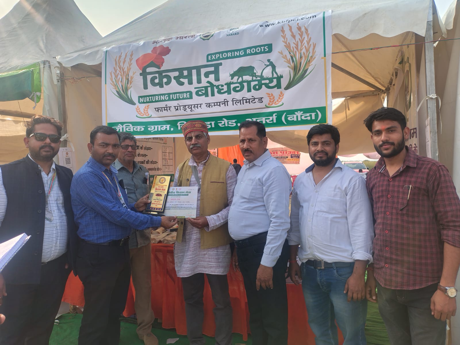 Officials presenting a trophy and certificate to a farmer at the Kisan Bodhgamya stall during a regional Kisan Mela at BUAT Banda